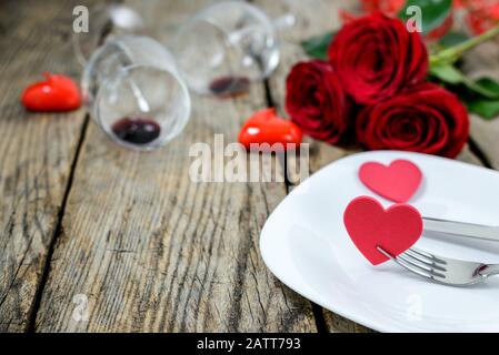 Herzen auf Platte mit Gabel und Messer, zwei Weinglas, rote Rosen und Kerzen auf einem alten Holztisch. Selektiver Fokus. Stockfoto