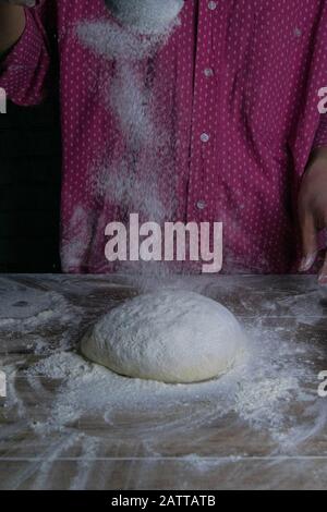 Die Frau bestreut Mehlteig aus der Fülle zum sieben von Mehl, Mehl fällt auf den Teig, eine schöne Streuung von Mehl. Bereiten Sie das Brot zu Stockfoto