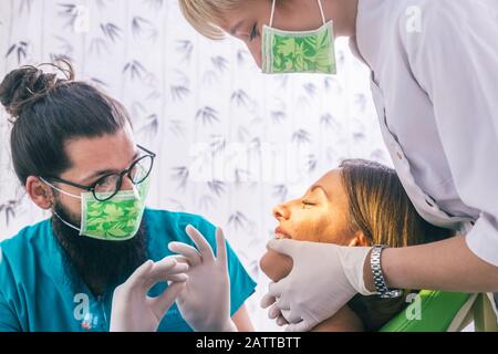 Nahaufnahme des Porträts einer schönen jungen Frau, die im Zahnstuhl sitzt, während die Stomatologin (Zahnärztin) mit den Händen in sterilen Handschuhen ihr T-Shirt befestigt Stockfoto
