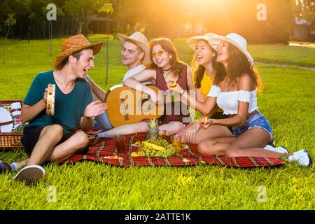 Gruppe junger Freunde Musiker auf Picknickparty, zusammen auf rotem Teppich im Park sitzen, Gitarre spielen und lachen. Stockfoto