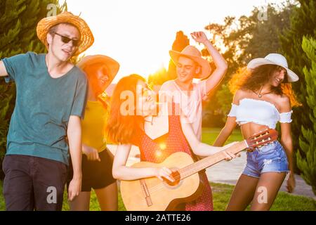 Gruppe glücklicher junger Freunde, die singen und tanzen und den Sommer im Park genießen. Halblanges Portrait eines Mädchens, das Gitarre spielt, tanzt und lächelt. Stockfoto