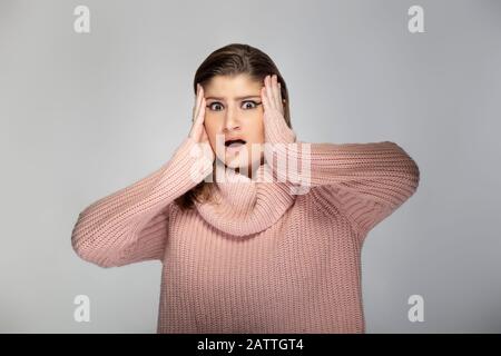 Nahaufnahme des Porträts einer jungen kaukasischen Frau, die einen rosafarbenen Pullover auf grauem Hintergrund trägt. Das Modell sieht schockiert und erschrocken aus Stockfoto
