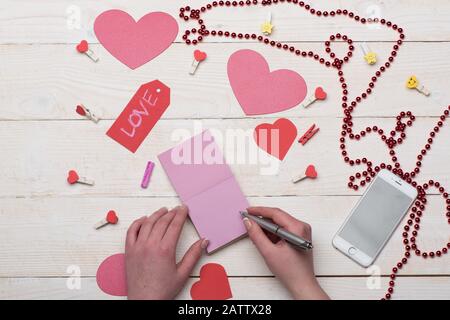 Valentinskarten, Dekorationen und Handy, Draufsicht. Weibliche Hand auf pinkfarbenem Notizpapier. Papierherzen, rote Perlen und niedliche Perlen auf weißem Vintage-Hintergrund. Liebe und Urlaubskonzept. Stockfoto