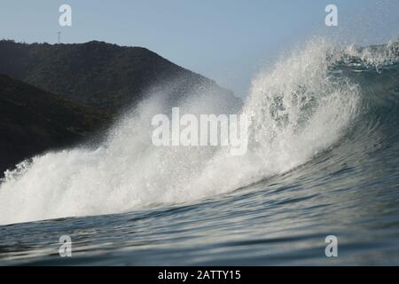 Eine große brechende Welle mit Hügeln und Tasman-Aussichtspunkt im Hintergrund am Piha Beach, Auckland, Neuseeland. Stockfoto