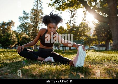 Portrait einer entschlossenen Fitness jungen afroamerikanischen Frau, die auf grünem Gras sitzt und im Park Sport macht - junge schwarze Frau, die sie erwärmt Stockfoto