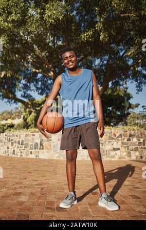 Aktives lächelndes Porträt eines Basketballspielers, der vor dem Baum im Park steht Stockfoto