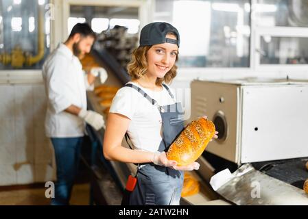 Porträt eines Bäckers mit Brot in den Händen vor dem Hintergrund einer automatisierten Linie in einer Bäckerei. Industrielle Brotproduktion Stockfoto