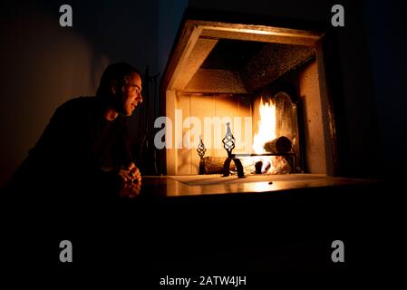 Mann sitzt vor einem brennenden Feuer auf einem offenen Kamin oder Kamin, der sich nach vorne neigt, um in die Flammen zu starren, als wäre er fasziniert Stockfoto