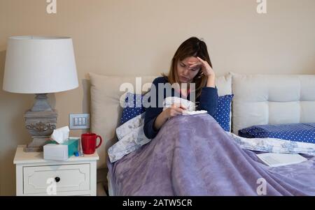 Kranke Frau, die ihre Temperatur an einem Thermometer überprüft, um zu sehen, ob sie Fieber hat, während sie zuhause im Bett ruht Stockfoto