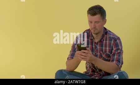 Fröhlicher Mann in kariertem Hemd auf dem Boden sitzend und auf Smartphone plaudern. Es ist ein gelber Hintergrund vorhanden. Stockfoto
