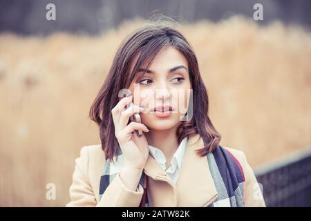 Frau in einem Telefongespräch. Mädchen im Herbst Beige Mantel blau weiß Schal sprechen auf Handy isoliert draußen hellbraunen Hintergrund Stockfoto