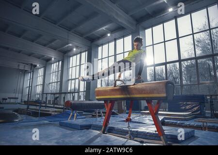 Champion. Kleine männliche Turnerin im Fitnessstudio, flexibel und aktiv. Kaukasischer Junge, Sportler in Sportswear, der in Übungen für Stärke und Balance praktiziert. Bewegung, Action, Bewegung, dynamisches Konzept. Stockfoto