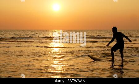 Silhouette des fröhlichen Surfmannes mit langen Surfbrettern bei Sonnenuntergang am tropischen Strand. Surfer am Strand an der Küste bei Sonnenuntergang mit schöner Aussicht Stockfoto