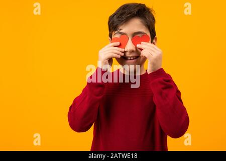 Lustiger fröhlicher gutaussehender kaukasischer Junge mit Papierkarten in Form von Herzen für Valentinstag auf orangefarbenem Hintergrund Stockfoto