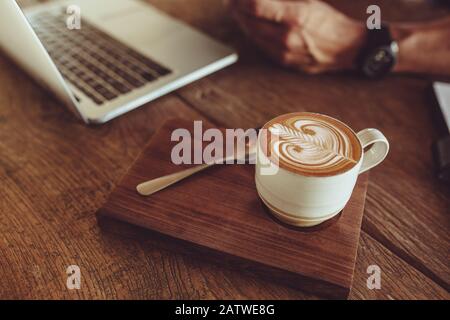 Nahaufnahme einer Tasse Cappuccino-Kunstkaffee auf dem Café-Tisch mit einem Mann, der am Laptop arbeitet. Kaffeetasse mit Kunstmuster aus Milchschaum. Stockfoto