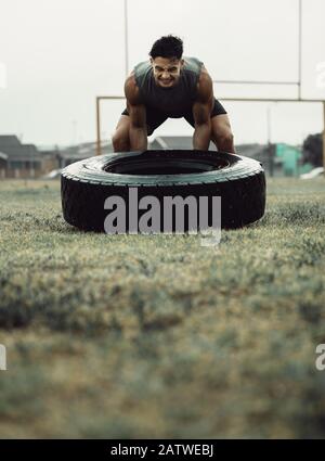 Starker junger Mann, der ein Flip-Workout für Reifen macht. Starker und muskulöser Athlet macht Cross-Training auf einem Feld. Stockfoto