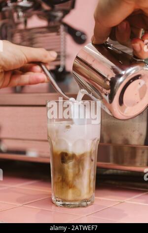 Geben Sie kochende Milch mit der Hand in ein Glas Eiskaffee, um Latte mit einem Löffel zu machen Stockfoto
