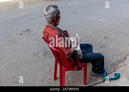 Ein armer 75 Jahre alter Asiat, der ein Schlaganfallopfer ist, sitzt in einem roten Plastikstuhl auf einer Stadtstraße im städtischen Kampong Cham, Kambodscha. Stockfoto