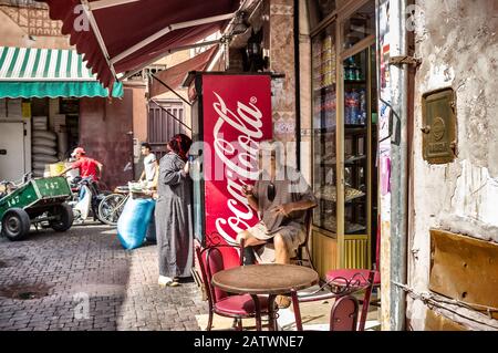Marrakesch, Marokko. Oktober 2015. Der alte Mann sitzt in traditioneller marokkanischer Kleidung außerhalb eines Cafés, das Minztee neben einem modernen Coca Cola Machin trinkt Stockfoto