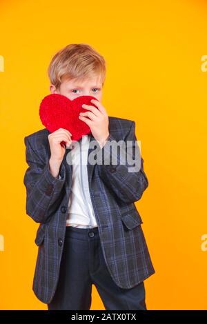 Kleiner blonder Junge, der Herz hält und mit der Kamera auf gelbem Hintergrund lachen kann. Stockfoto