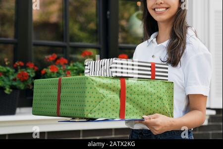 Seitenansicht des inkognito jungen Kuriers in weißem T-Shirt mit zwei Geschenkschachteln im Freien. Fröhliche brünette Frau in der Nähe des Hauses, hält Geschenke und Mappen und lächelt. Lieferkonzept. Stockfoto
