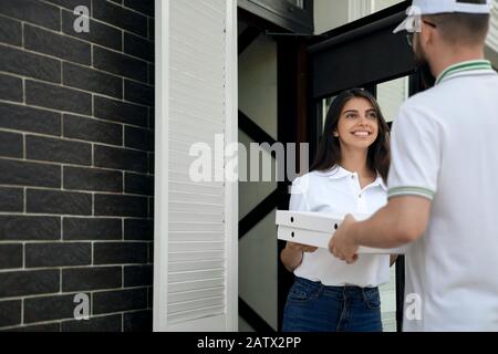 Rückansicht des jungen Zustelljungen in weißer Kappe und Brille, die zwei Kisten Pizza an die Frau gibt. Hübsche, lächelnde Brunette-Frau in legeren Outfits, die im Freien bestellt wird. Konzept für die Lieferung von Lebensmitteln. Stockfoto