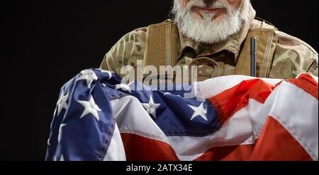 Nahaufnahme des bärtigen amerikanischen Militärveteranen, der die Flagge in Waffen hält. Porträt von inkognito altem stolzen männlichen Offizier in Tarnuniform, der in dunkler Atmosphäre posiert. Begriff des Militärs, Patriotismus. Stockfoto