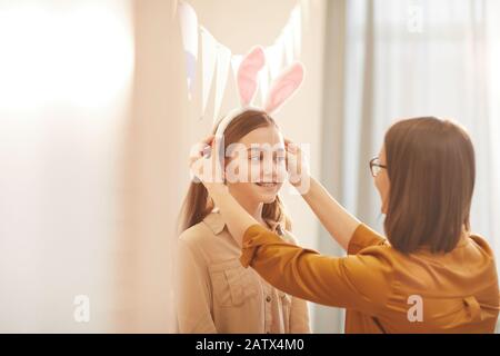 Mutter, die Kaninchenohren auf dem Kopf ihrer Tochter trägt, bereitet sie Kostüm für den Osterfeiertag vor Stockfoto