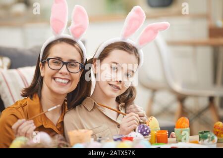 Porträt von glücklicher Mutter und Tochter in Kaninchenohren lächelnd vor der Kamera, während sie mit Farben am Tisch sitzen Stockfoto