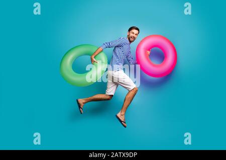 Lange Foto von funky verrückten Kerl Tourist Jump hoch Laufschwimmen halten zwei bunte Rettungsschwimmerkreise tragen gestreifte Matrosen Hemd Shorts Flip Stockfoto