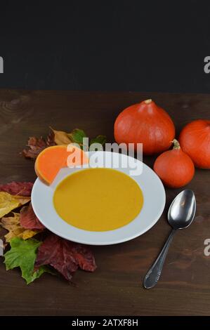 Herbstliche Kürbissuppe in einer Schüssel mit hokkaido-kürbissen, Blättern und einem Löffel auf einem braunen Holztisch Stockfoto