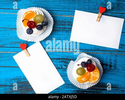 Weißes viereckiges Blatt Papier wird von roten Holzkleidern in Form von Herzen auf trendiger blauer Holzoberfläche mit köstlichem pavlova-kuchen befestigt Stockfoto