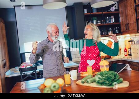 Das schöne Senioren-Paar tanzt und lächelt beim gemeinsamen Kochen in der Küche Stockfoto
