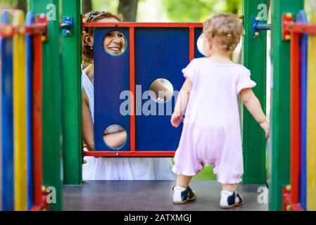 Fröhliche Mutter, die ihr kleines Mädchen auf dem Spielplatz betrachtet. Einjähriges Kind spielt mit der Mutter. Stockfoto