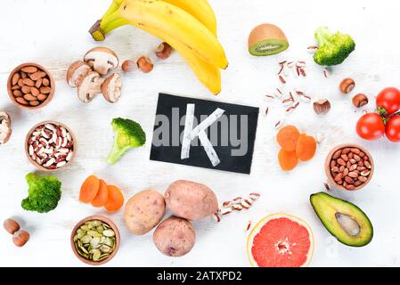 Lebensmittel, die natürliches Kalium enthalten. K: Kartoffeln, Pilze, Banane, Tomaten, Nüsse, Bohnen, Brokkoli, Avocados. Draufsicht. Auf weißem Holzhintergrund. Stockfoto