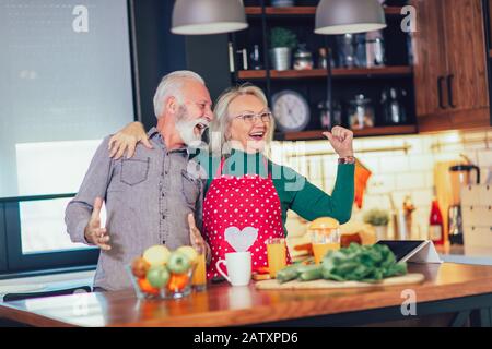 Das schöne Senioren-Paar tanzt und lächelt beim gemeinsamen Kochen in der Küche Stockfoto