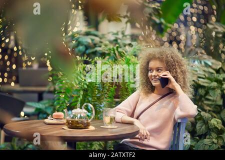 Junge lächelnde Frau mit lockigen Haaren, die im Café mit einer Tasse Tee und einem Telefongespräch ruht Stockfoto
