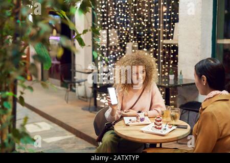 Junge Frau, die ihr Handy hält und ihrem Freund während ihres Mittags im Café Fotos zeigt Stockfoto