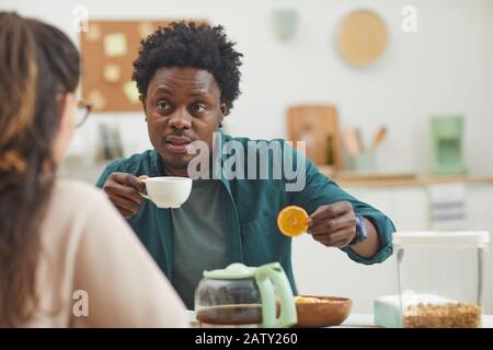 Afrikanischer junger Mann trinkt Kaffee mit Keksen und spricht mit seiner Freundin in der Küche zu Hause Stockfoto