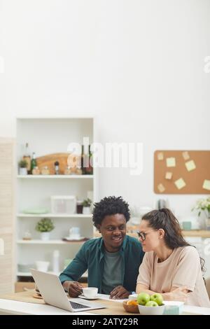 Multiethnisches junges Paar, das zusammen frühstücken und einen Laptop benutzen, während es zu Hause in der Küche sitzt Stockfoto
