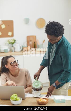 Afrikanischer junger Mann, der Kaffee in die Tasse seiner Freundin gießt, während sie am Laptop am Tisch in der Küche arbeitet Stockfoto