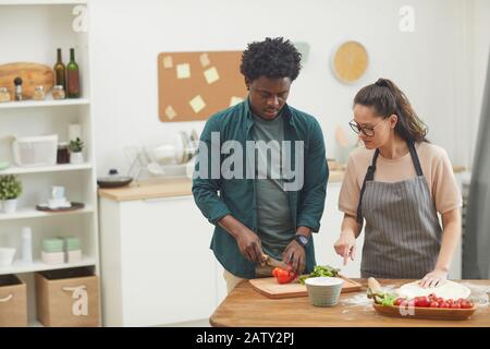 Afrikanischer junger Mann, der mit seiner Frau, die Teig zubereitete, Gemüse auf dem Schneidebrett schneidet, den sie in der Küche zu Hause kochen Stockfoto