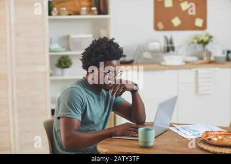 Afrikanischer junger Mann, der am Tisch sitzt und in der Küche zu Hause am Laptop arbeitet Stockfoto