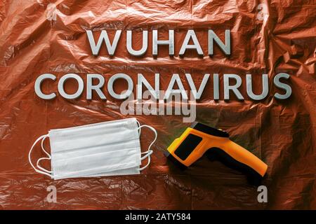 Worte wuhan Coronavirus mit Metallbuchstaben auf orange zerknitterter Kunststoff-Filmkulisse mit Gesichtsmasken und Infrarotthermometer - flach mit dram Stockfoto