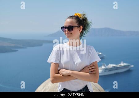 Junge, schöne Jugendliche, Mädchen, die lächelnd posieren, Hintergrund Sommer sonnige Landschaft am Meer mit weißen Kreuzfahrtschiffen in der ägeischen See auf der berühmten Gr Stockfoto