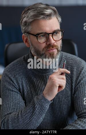 Erwachsener Geschäftsmann im Büro. Nahbetriebsportrait des gutaussehenden grauen Haares, der Brille trägt, die am Arbeitsplatz sitzt. Selbstbewusster Geschäftsmann winkt Stockfoto