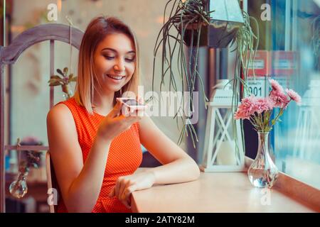 Portrait eines Mädchens mit der Spracherkennung des Telefons und Blick auf das Handy in der Nähe eines Fensters eines Hauses oder einer Café-Bar Stockfoto