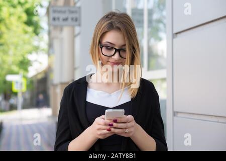 Eine glückliche Dame, die mit einem Smartphone Online-Inhalte auf der Straße beobachtet, lächelt in Geschäftskleidung und schwarzer Brille. Gemischtes Rennmodell Stockfoto