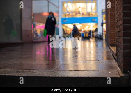 Unscharf Bild einer Frau, die im Stadtzentrum mit Lichtreflexen und Ladenfronten auf einem verschwommenen nassen Bürgersteig einkauft Stockfoto