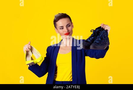 Liebe zum bequemen Schuhkonzept. Frau kurze Haare Mädchen mit gelben High Heels Schuhfarbe in einer Hand Blick auf die flachen sportlichen Schuhe blau Stockfoto